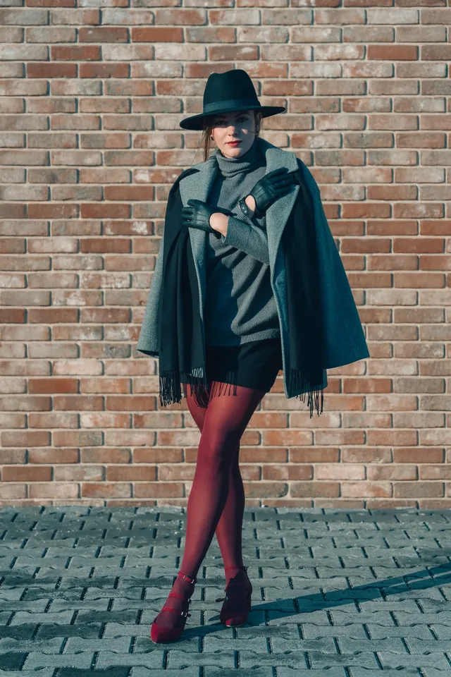 Full-length portrait of a woman in a hat and red trousers beside a wall.