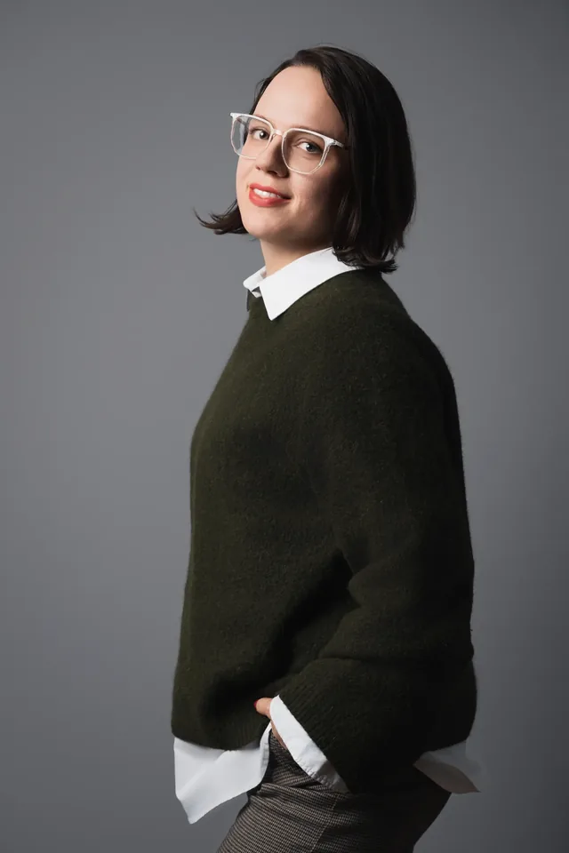 Woman wearing glasses standing sideways against a grey background.