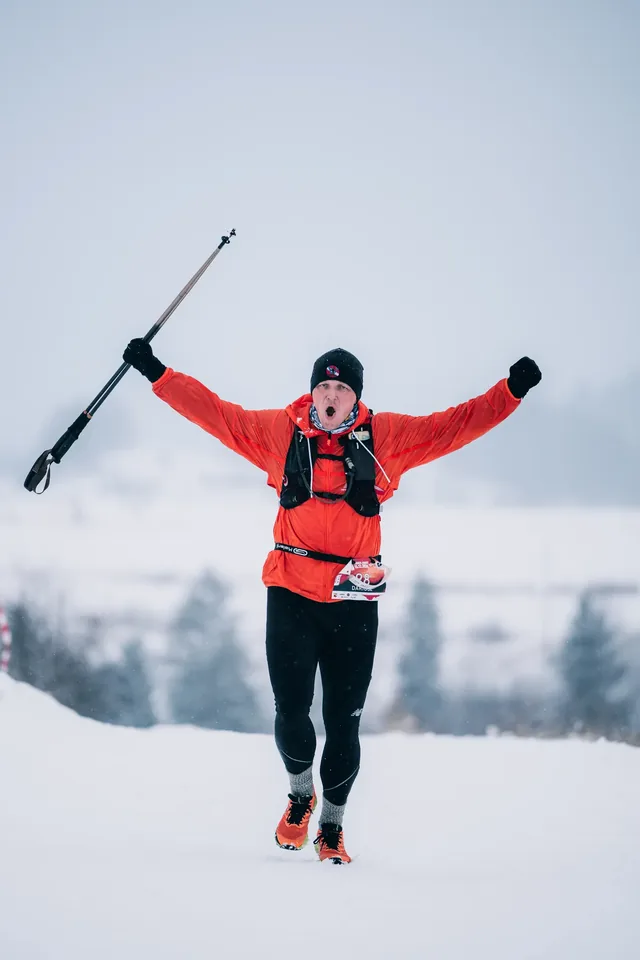 Runner in a red jacket with raised arms and a pole.